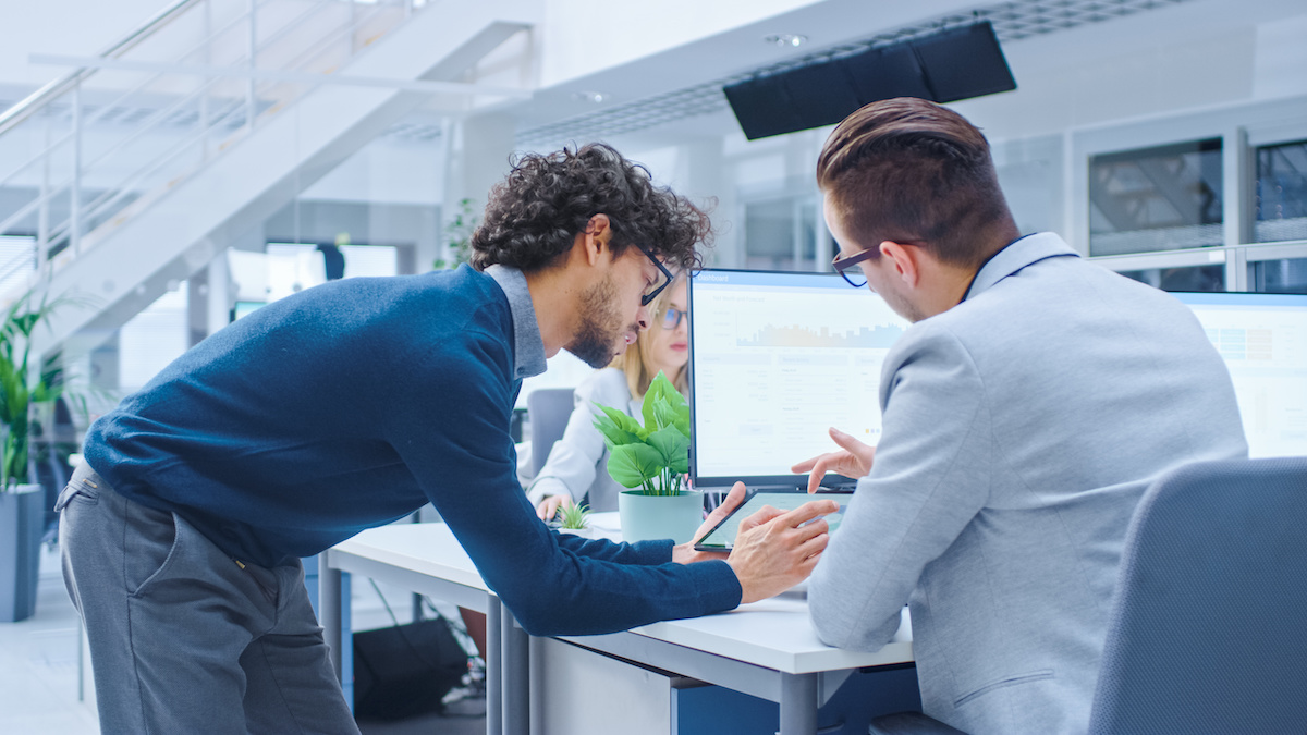 In Bright Modern Office: Office Worker Sitting and Working at His Desktop Computer, Discusses Problem Solution with His Project Manager and Team Leader. Young Businessmen Planning and Problem Solving